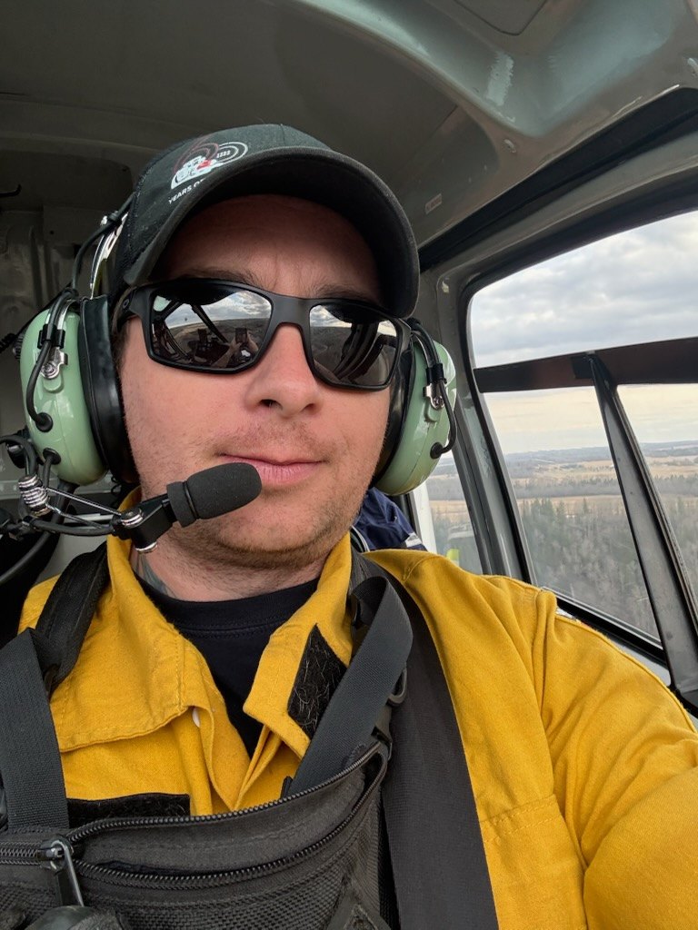 Sean Cunningham in a helicopter wearing a headset and a yellow wildland fire shirt, flying over forest