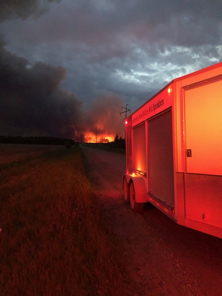 A wildfire burning on the horizon at dusk, with a structure protection sprinkler trailer staged on a rural road